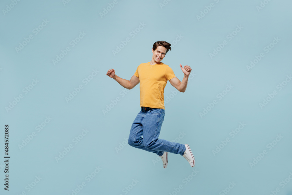 Full body overjoyed excited fun cool young man 20s wear yellow t-shirt jump high do winner gesture clench fist isolated on plain pastel light blue background studio portrait. People lifestyle concept.