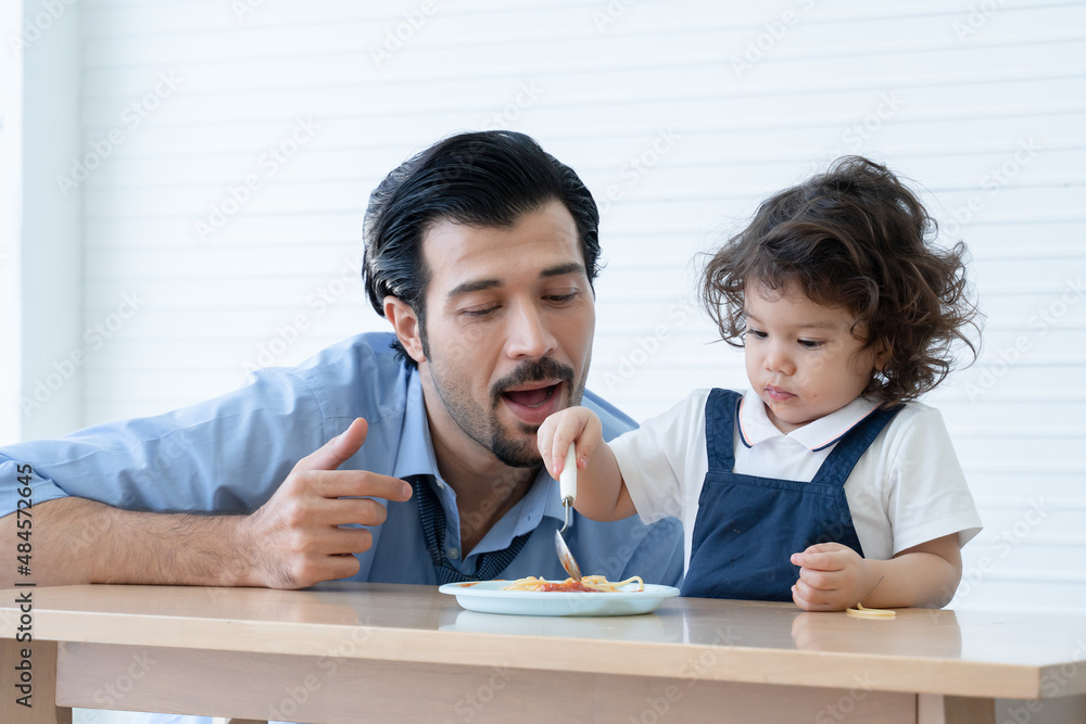 Caucasian young father with beard looking and cheer up little cute daughter trying to eat spaghetti with spoon by herself at home and adorable kid girl enjoy eating with face is mess up with ketchup Caucasian young father with beard looking and cheer up little cute daughter trying to eat spaghetti with spoon by herself at home and adorable kid girl enjoy eating with face is mess up with ketchup