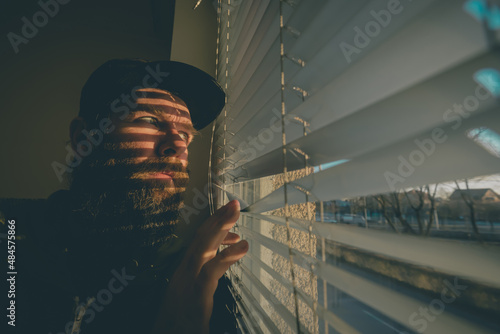Young hipster male with beard looking through blinds as if he is spying on someone. Suspicious male looking through sun lit venetian blinds.
