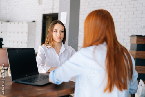 Back view of two serious focused young business women discussing new project sharing ideas sitting at desk opposite each other in office meeting room. Concept of job interview human resources.