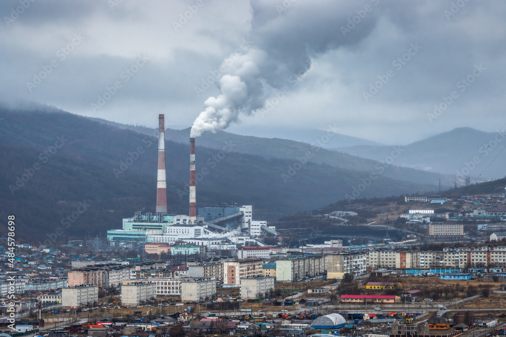 Urban industrial landscape. Top view of residential buildings and thermal power plant. Thick puffs of smoke from the factory chimney. Cloudy rainy weather. Magadan, Magadan region, Siberia, Russia.