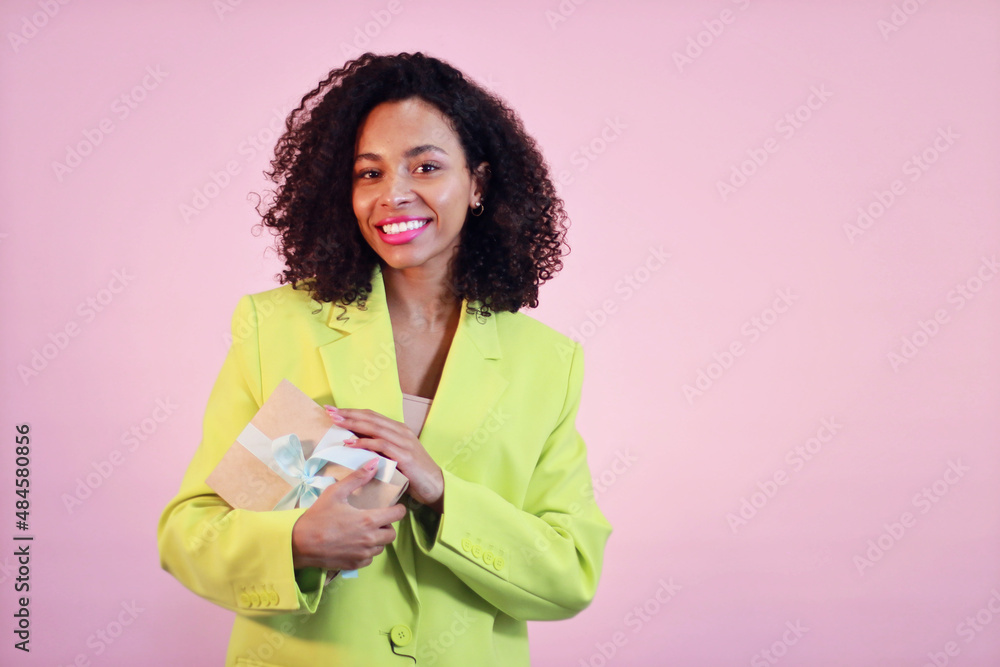 Smiling and young african american women on pink background in lime jacket and present box. Smiling and young african american women on pink background in lime jacket and present box.