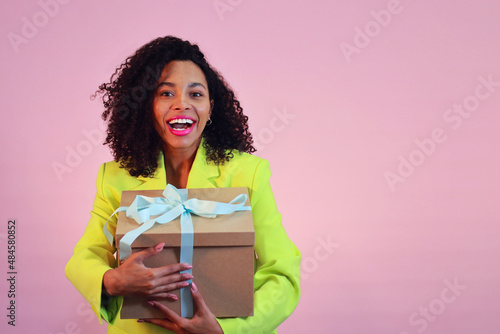 Smiling and young african american women on pink background in lime jacket and present box.