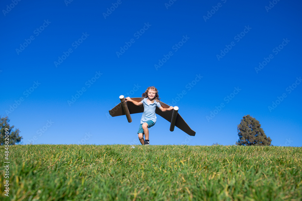 Happy child playing outside on green grass and blue sky. Kid pilot with toy jetpack. Kid boy play with toy plane cardboard. Summer travel and adventure. Success winner, imagination and dream concept. Happy child playing outside on green grass and blue sky. Kid pilot with toy jetpack. Kid boy play with toy plane cardboard. Summer travel and adventure. Success winner, imagination and dream concept.