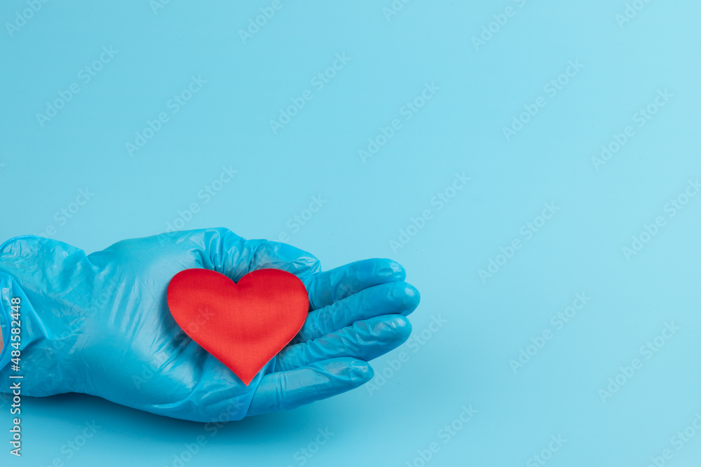 Fototapeta premium Red heart in hands in blue medical gloves on blue background. Background for the day of the medic. World Heart Day. Heart diseases.