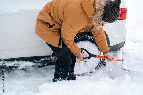 A man digs out a stalled car in the snow with a car shovel. Transport in winter got stuck in a snowdrift after a snowfall, sat on the bottom. First aid, tow truck, winter tires spikes and all-season