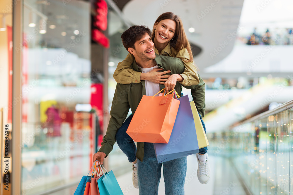 Spouses Shopping Having Fun, Husband Carrying Wife Piggyback In Hypermarket Spouses Shopping Having Fun, Husband Carrying Wife Piggyback In Hypermarket