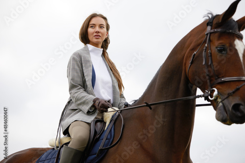 Beautiful young woman riding a horse outdoor. Concept of animal care. Rural rest and leisure. Idea of green tourism. Young european woman wearing helmet and uniform. © Georgii
