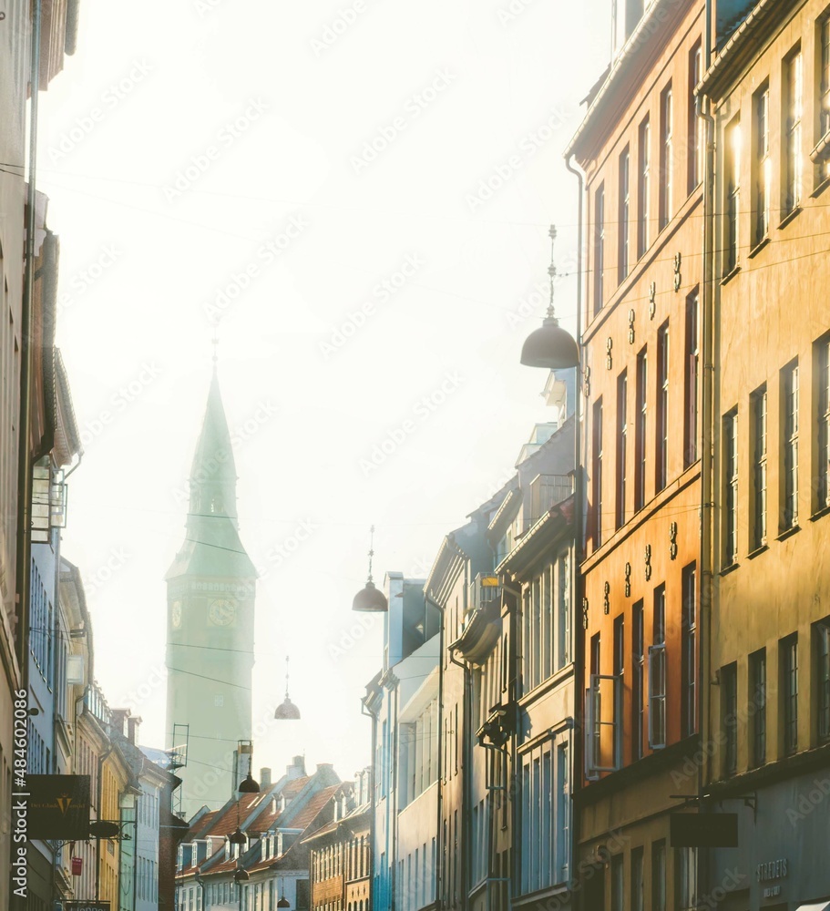 Fototapeta premium Street in the inner city of Copenhagen with a silhouette of the iconic clocktower of the city hall in the background. overexposed bright sunlight.