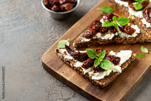 Bruschetta or whole grain bread sandwich with cream cheese and sun-dried tomatoes. Crostini on a wooden serving board on a dark kitchen table closeup 