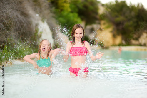Two young sisters bathing in natural swimming pool in Bagno Vignoni, with thermal spring water and waterfall. Tuscany, Italy.