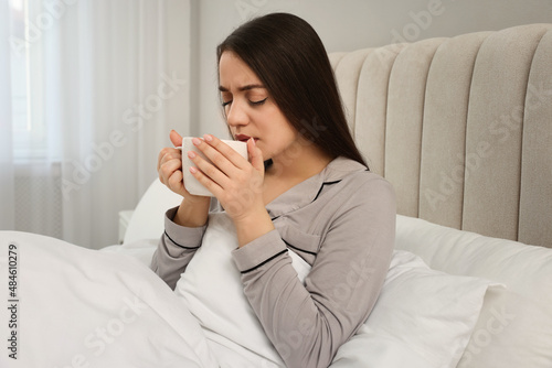 Sick young woman with cup of hot drink in bed at home