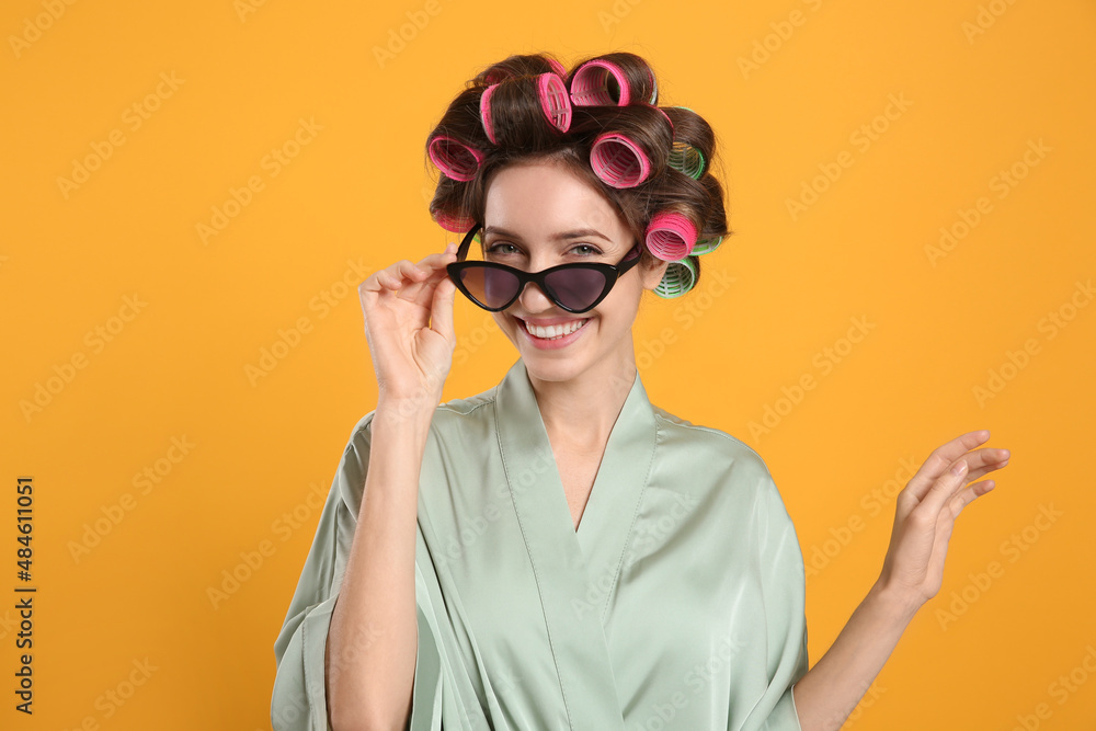 Beautiful young woman in silk bathrobe with hair curlers and sunglasses on yellow background Beautiful young woman in silk bathrobe with hair curlers and sunglasses on yellow background