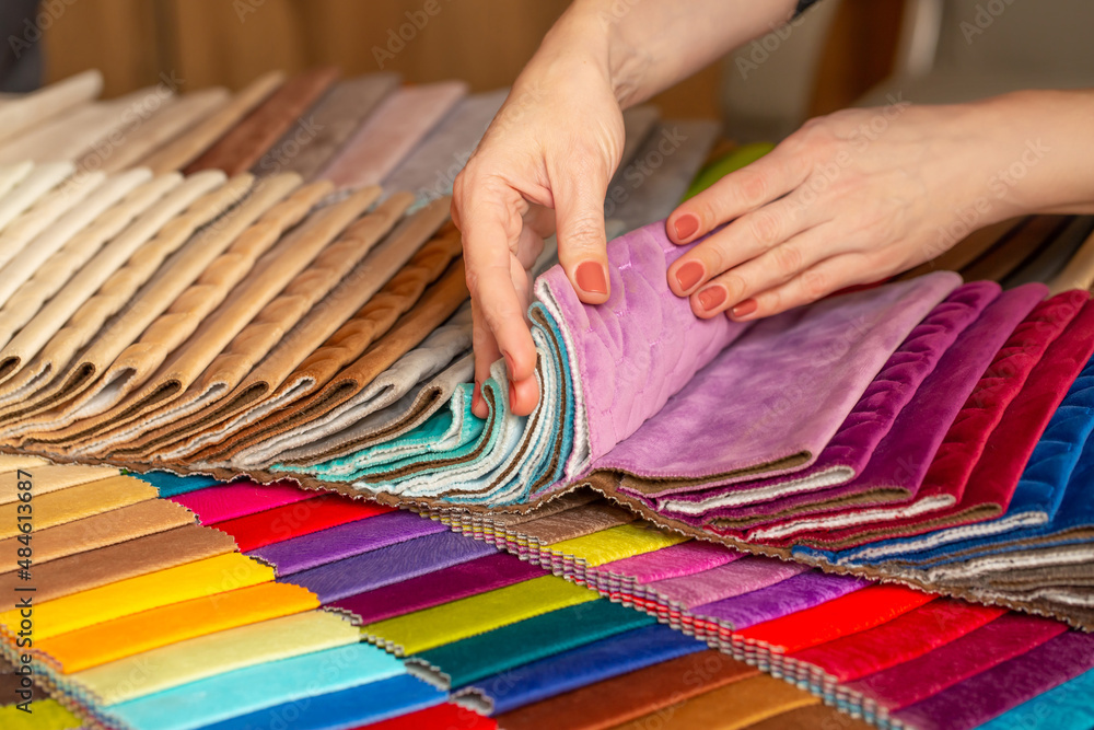 Fabric swatches in different colors are stacked for selection. A woman chooses upholstery colors for furniture and interiors. Touches the texture of the fabric, hands on the material close-up.