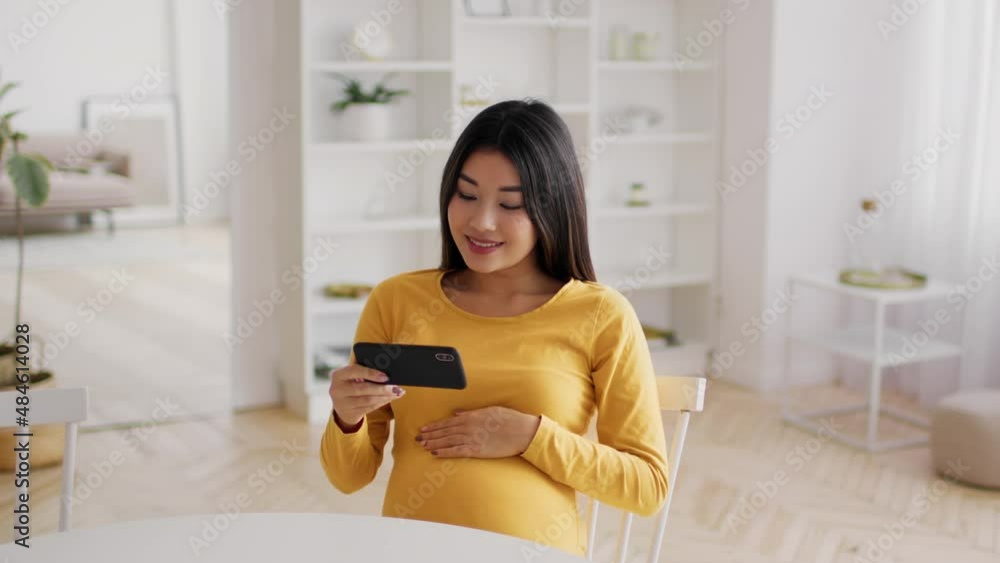 Young pregnant asian woman watching videos on smartphone while relaxing at home Young pregnant asian woman watching videos on smartphone while relaxing at home
