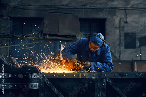 Fototapete Caucasian man in special overalls, gloves and glasses using angle grinder for polishing metal construction at workshop