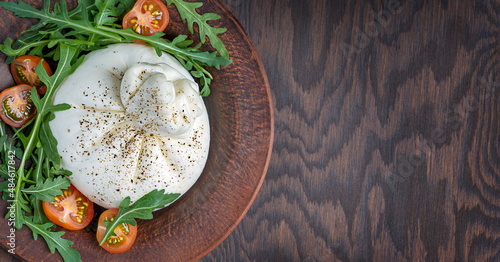 Top view of italian burrata white homemade soft milk cheese made of mozzarella and cream served with sliced cherry tomatoes and arugula leaves o plate on dark brown wooden background with copy space