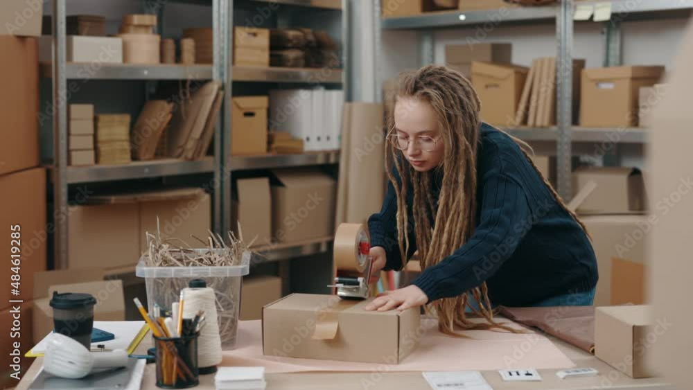 Young female online store owner, warehouse worker, merchant seller seals with tape package, preparing delivery parcel box. Post service and small business concept. Young female online store owner, warehouse worker, merchant seller seals with tape package, preparing delivery parcel box. Post service and small business concept.