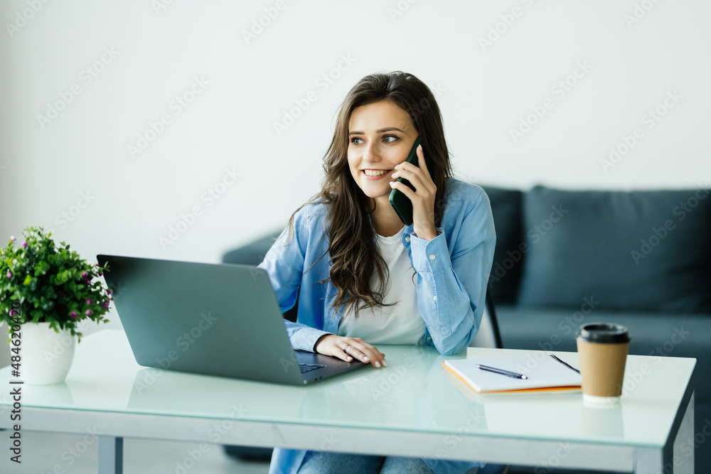 Attractive smiling young business womantalking on mobile phone while sitting on a desk and using laptop computer in office Attractive smiling young business womantalking on mobile phone while sitting on a desk and using laptop computer in office