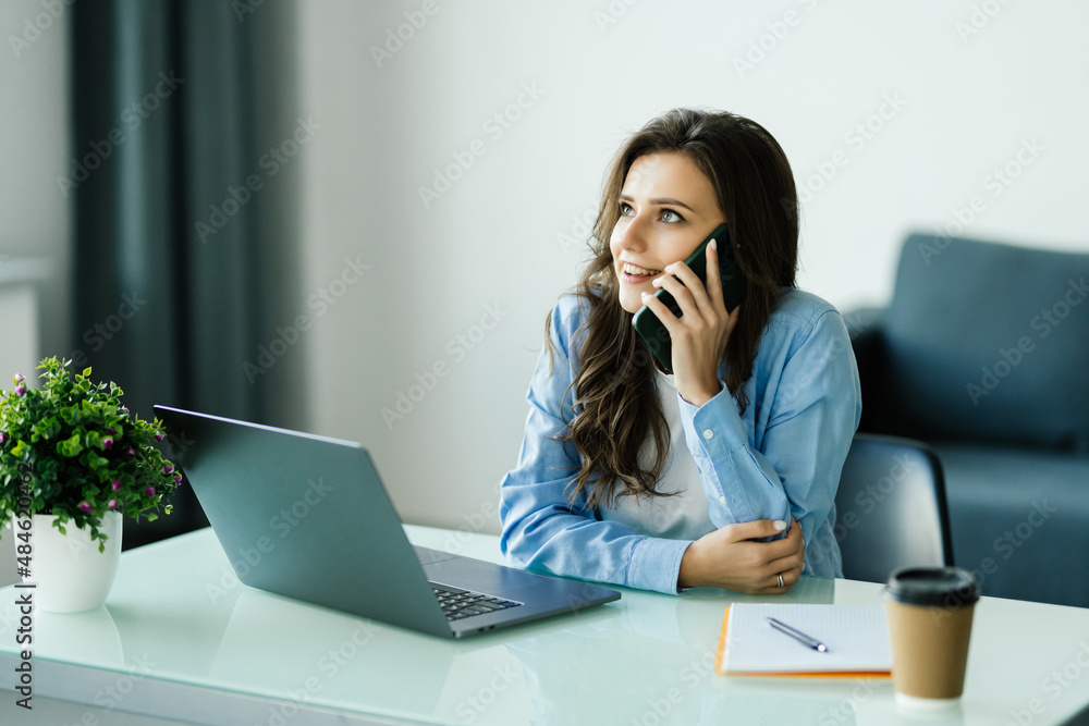 Attractive smiling young business womantalking on mobile phone while sitting on a desk and using laptop computer in office Attractive smiling young business womantalking on mobile phone while sitting on a desk and using laptop computer in office