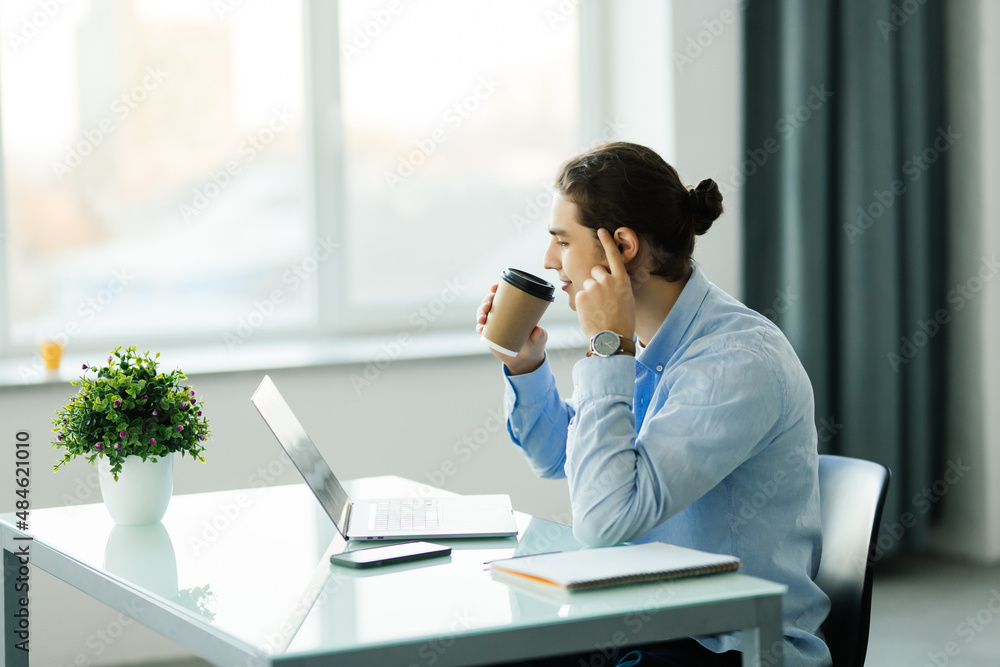Young businessman using computer and drinking coffee in office. Handsome man on coffee break Young businessman using computer and drinking coffee in office. Handsome man on coffee break