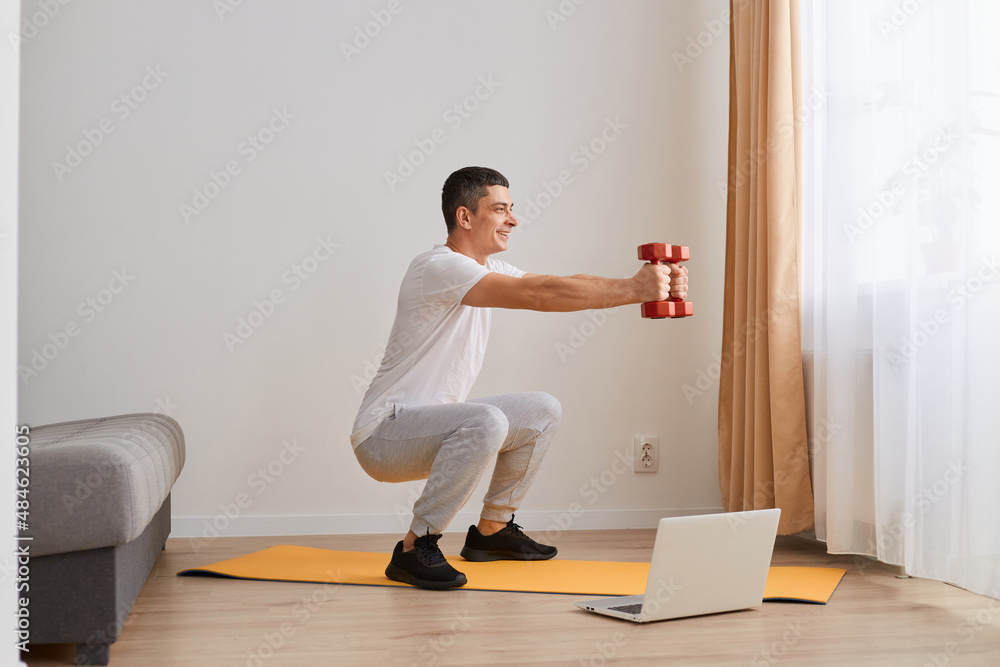 Full length shot of male fitness man squatting with dumbbells via video lesson on training at home, using laptop, sporty guy wearing white t shirt and gray pants. Full length shot of male fitness man squatting with dumbbells via video lesson on training at home, using laptop, sporty guy wearing white t shirt and gray pants.