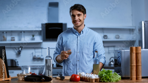 happy young man holding glass of white wine near table with fresh ingredients.