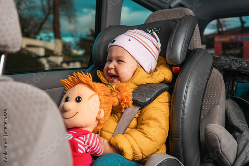 Portrait of happy smiling caucasian little child is sitting strapped in a child seat with a plush toy in his hands. View inside a car