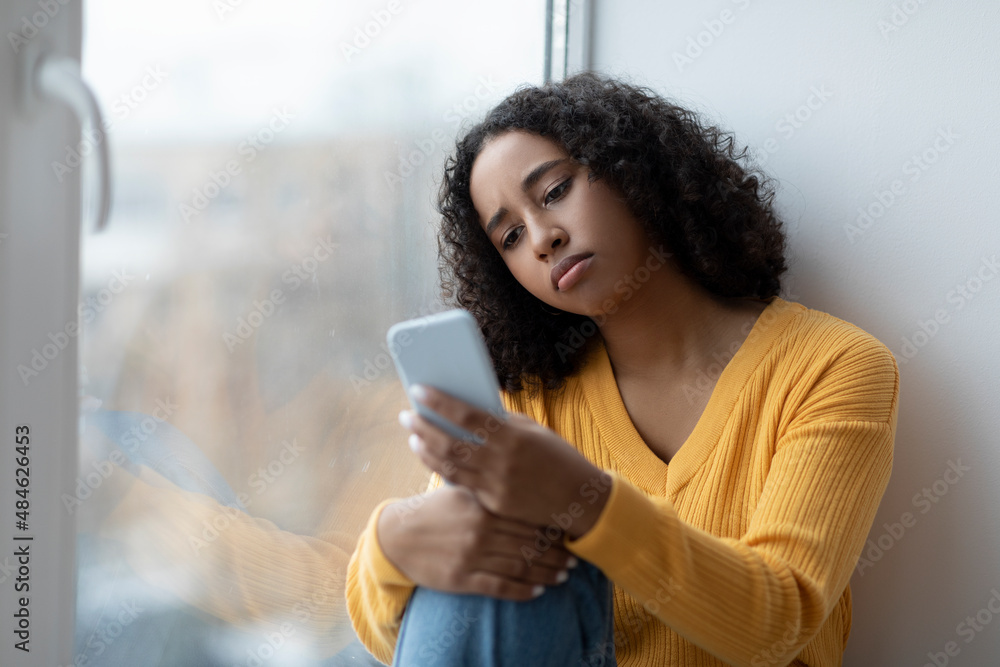 Depressed young African American woman reading upsetting news on smartphone, sitting near window at home, copy space