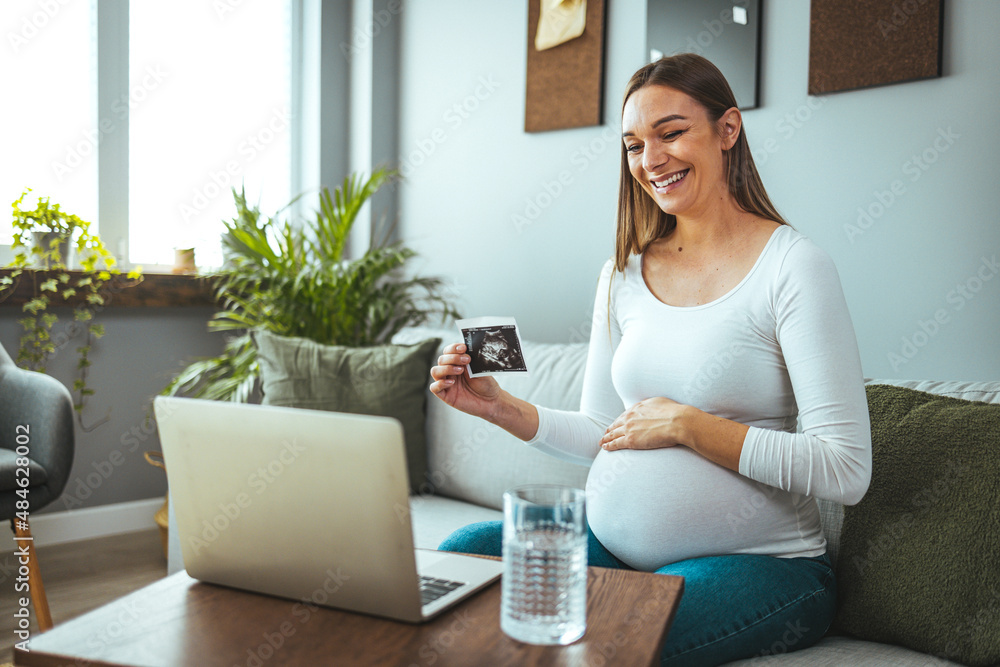Pregnancy, motherhood, concept - happy pregnant woman with laptop who has a video call and shows an ultrasound image with a toothy smile. Pregnancy, motherhood, concept - happy pregnant woman with laptop who has a video call and shows an ultrasound image with a toothy smile.