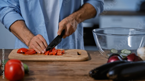 cropped view of man cutting tomatoes near blurred eggplants and glass bowl.