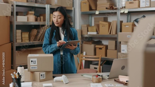 Waist up portrait view of the asian girl holding tablet computer and checking information about parcel on it while preparing sending it to the client. Small business concept.