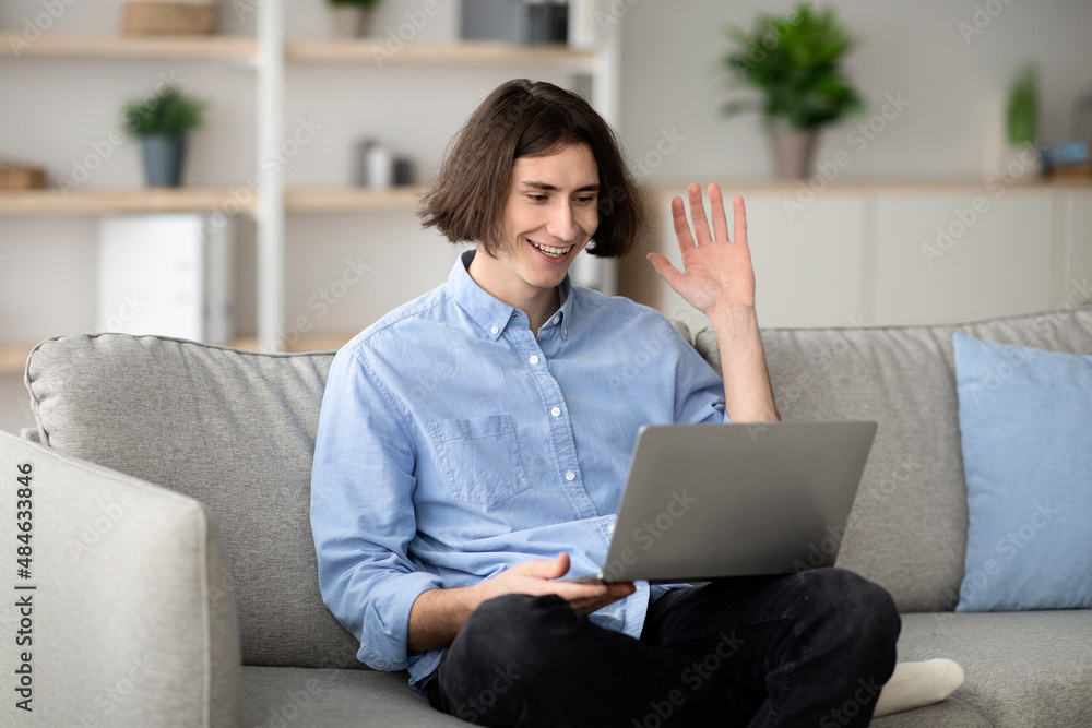 Distant communication concept. Happy man video calling on laptop computer, waving hand to screen, sitting on sofa Distant communication concept. Happy man video calling on laptop computer, waving hand to screen, sitting on sofa