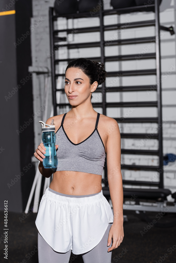 Smiling middle east sportswoman holding sports bottle in gym.