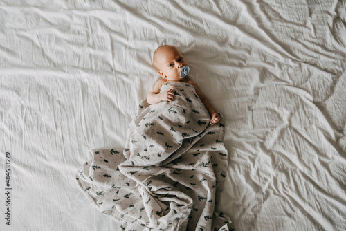 Εκτύπωση καμβά Newborn baby with a pacifier, covered with a muslin blanket, lying in bed on white bed sheet, top view