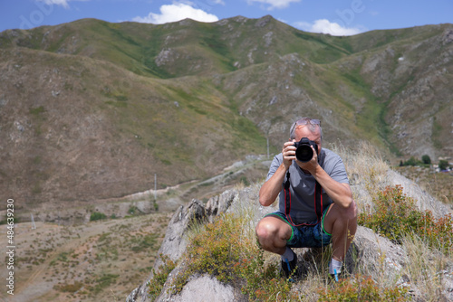 Man with a camera squatting on a grassy rock on the background of mountains.