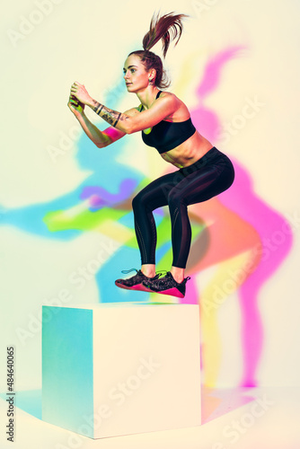 Sporty woman doing box jump exercise. Photo of athletic woman in black sportswear on white background with effect of rgb colors shadows. Strength and motivation. Full length. Side view