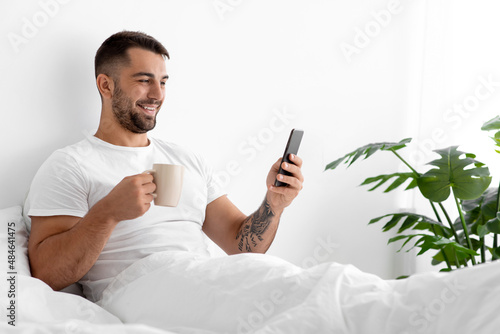 Happy caucasian attractive man in white t-shirt with cup of coffee in bed in bedroom #484641475