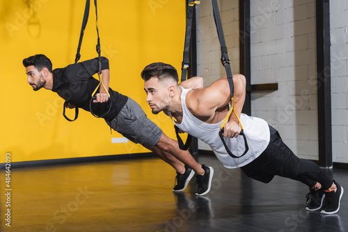 Young multiethnic sportsmen training with suspension straps in gym.