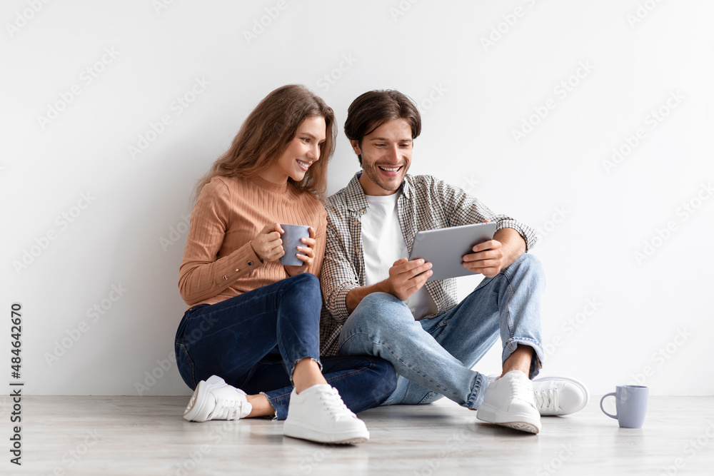 Satisfied millennial caucasian family sitting on floor planning new interior in tablet in empty room
