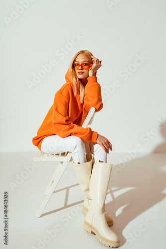 Fashionable confident blonde woman wearing trendy orange sweatshirt, white skinny jeans, high leather boots, posing on white background. Full-length stuido portrait