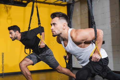 Athletic sportsman training with suspension straps near muslim man in gym.