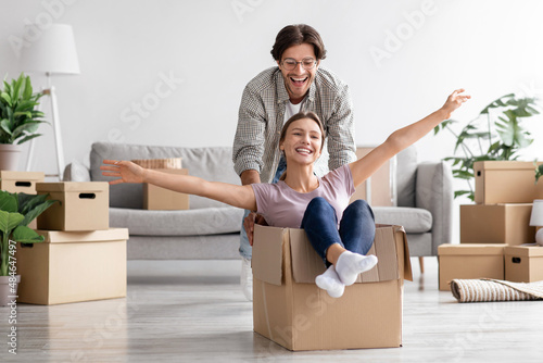 Fotografie Glad caucasian young man pushes wife in cardboard box have fun in living room in