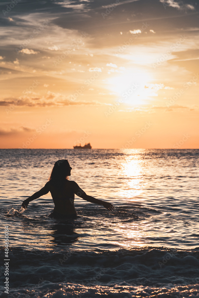 Silhouette of smiling woman on beach inside sea with her arms outstretched in front of sunset sun