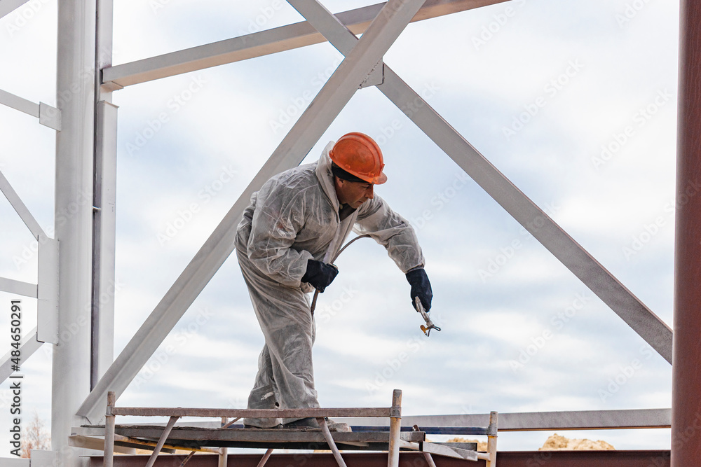 A worker in special protective clothing, works on painting and fire protection of metal structures at a height. Painting the metal at the construction site.