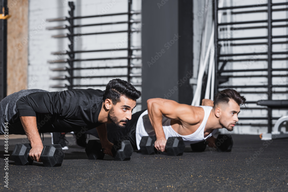 Naklejka premium Young arabian sportsman doing press ups near friend with dumbbells in gym.