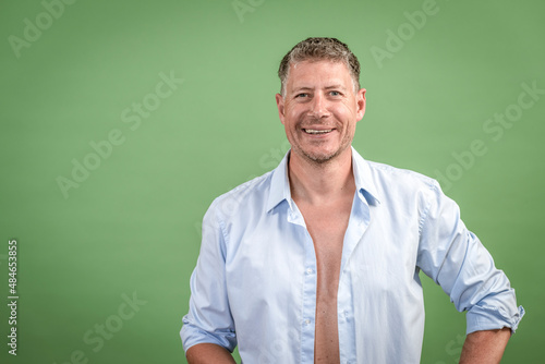Middle aged man with open blue shirt is standing in front of green wall