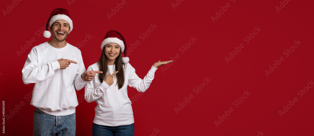 Fototapeta premium Holiday Promo. Cheerful Young Couple In Santa Hats Pointing At Copy Space