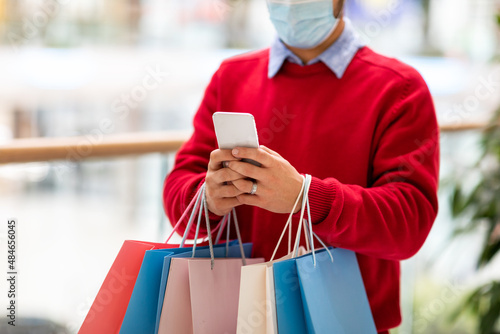 Cropped view of young man in face mask holding gift bags, using mobile shopping app on phone at city mall, closeup