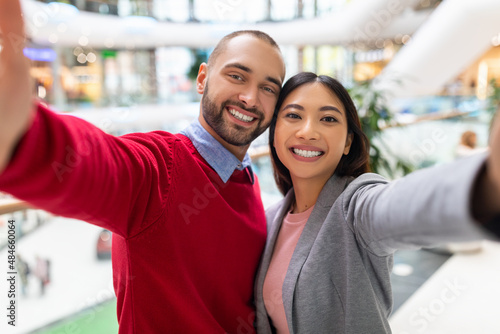 Loving young diverse couple smiling, taking selfie at huge shopping centre